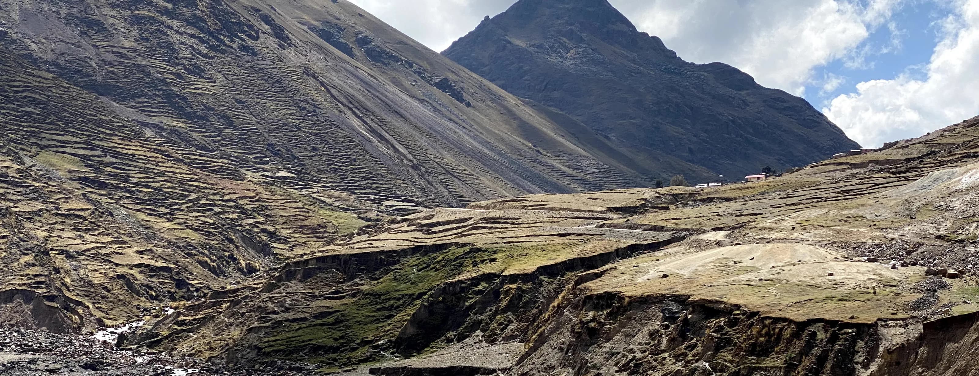 Valle andino entre laderas de pasto surcadas por hileras de tierra, con un cerro de roca al fondo.